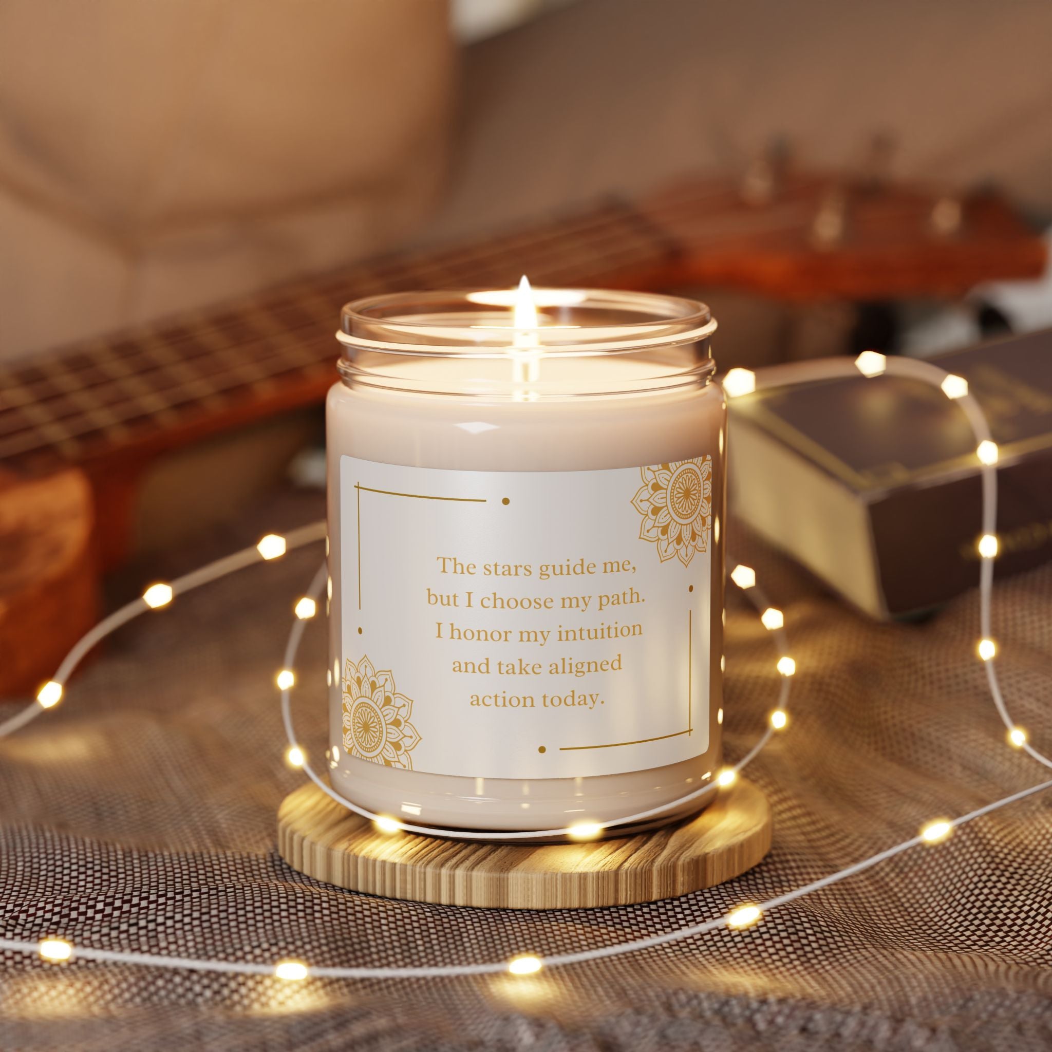 Candle with a motivational quote on a wooden coaster surrounded by string lights and a guitar in the background.
